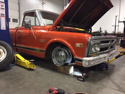 1984 Ford Bronco - An orange and white pickup truck is being worked on in a garage. The hood is open, and the front passenger side wheel is elevated on blocks…