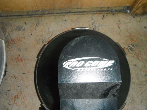 1990 Ford Bronco - A close-up view of a black Pro Comp branded air cleaner lid, likely for a Ford Bronco, resting on a dirty concrete floor next to a wooden w…