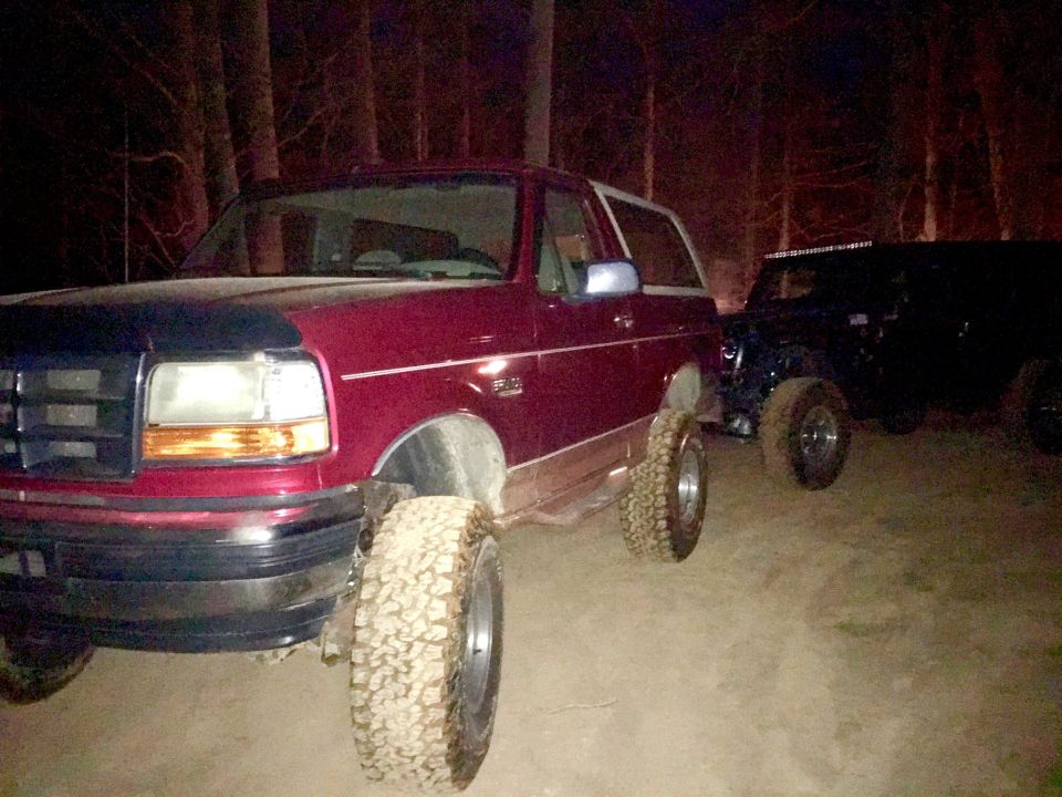 Maroon Ford Bronco with large off-road tires parked at night next to a Jeep.