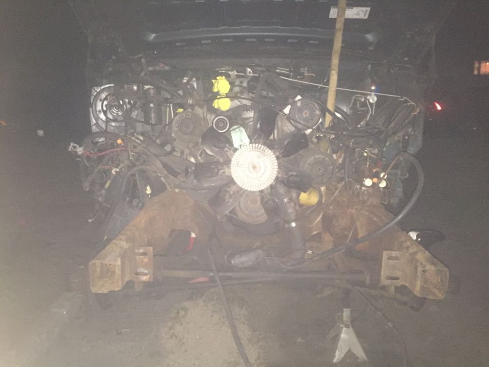 1995 Ford Bronco engine bay with hood open, partially disassembled engine, supported by jack stands outdoors at night.