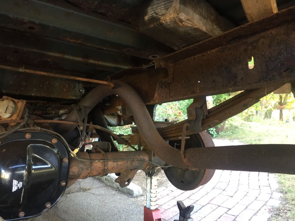 Underside of a rusty vintage vehicle showing frame, leaf springs, exhaust, and rear differential.