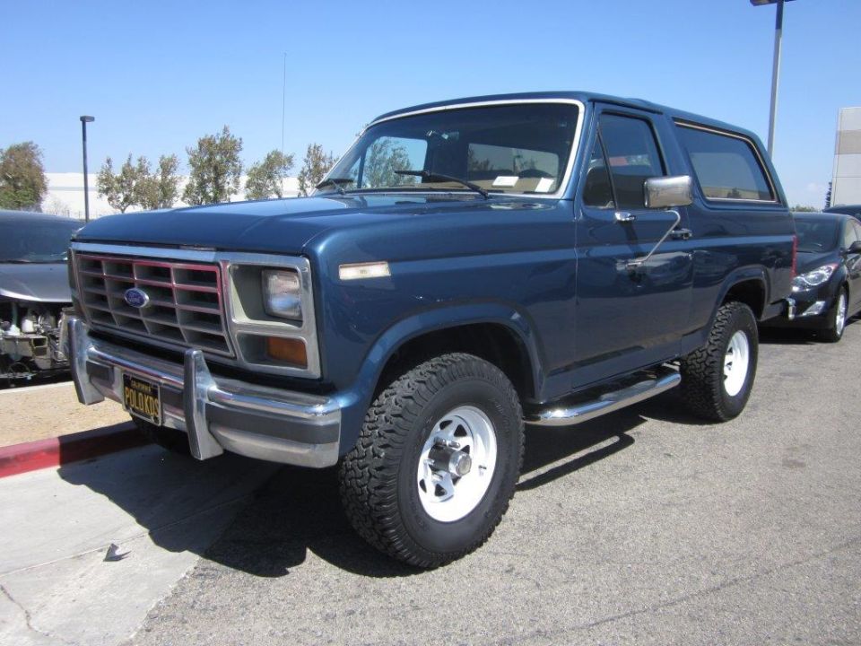 Blue 1980s Ford Bronco parked on asphalt lot, front 3/4 view.