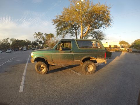 1996 Ford Bronco - A green Ford Bronco is parked in a lot with a large tree casting a shadow. Stickers are visible on the rear window.