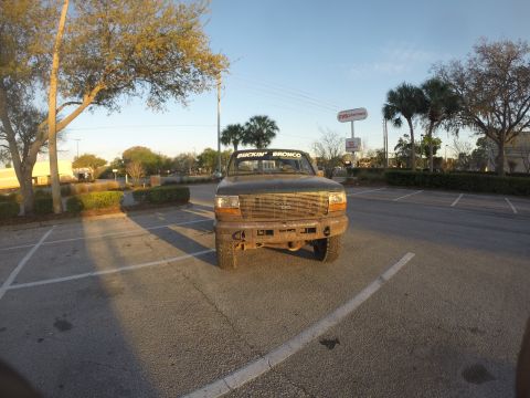 1996 Ford Bronco - A front-facing view of a dark green 1996 Ford Bronco parked in a parking lot during golden hour. The vehicle has a custom bumper and text o…