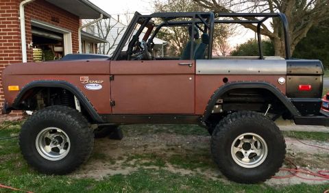 1972 Ford Bronco - The side profile of a modified, lifted 1972 Ford Bronco is shown in a driveway. The vehicle has a brown body, a black roll cage, and large…