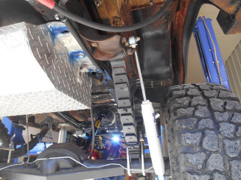1972 Ford Bronco - The underside of a vehicle, likely a 1972 Ford Bronco, shows a diamond plate fuel tank, leaf springs, and a shock absorber. A person is wel…