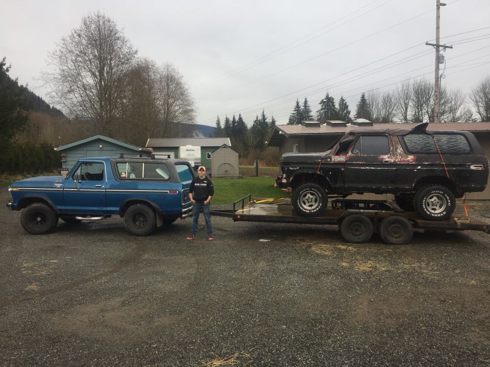 Blue 1978 Ford Bronco parked next to a black Ford Bronco on a trailer, person standing in between