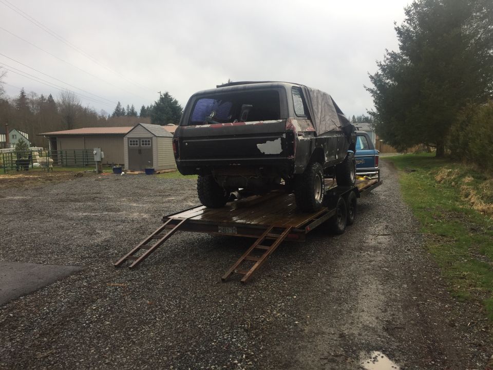 Black Ford Bronco on a flatbed trailer, covered with a tarp, parked on a gravel driveway.