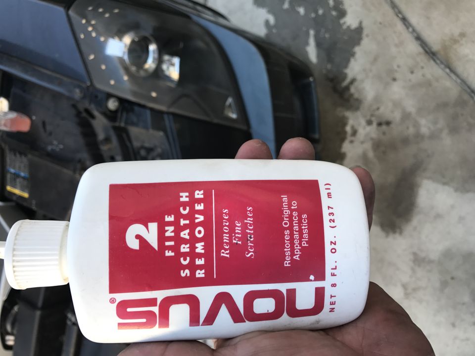 Hand holding Novus 2 Fine Scratch Remover bottle in front of a black car headlight.
