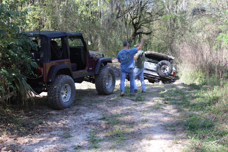 Two men pulling a white Ford Bronco out of a ditch with a rope, a red Jeep Wrangler is nearby.