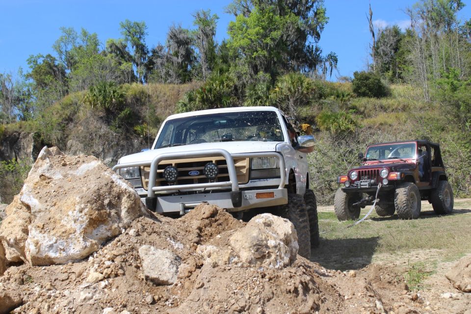 White 1992 Ford Bronco off-roading over rocks, red Jeep Wrangler in background.