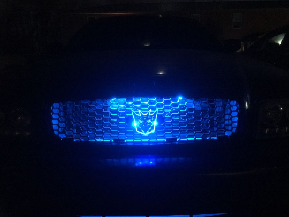 Dark sedan with blue LED-lit grille and Autobot logo at night.