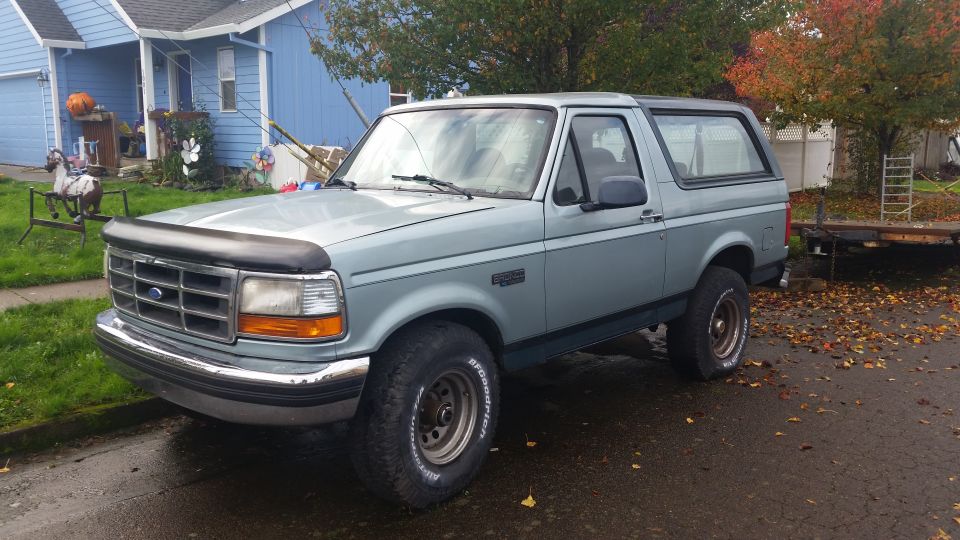 Light blue 1996 Ford Bronco parked on a wet driveway in front of a blue house, 3/4 front view.