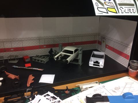 1989 Ford Bronco - A model car garage scene is set up with two unfinished model car bodies, one on a lift and another on the floor. Various model car parts an…