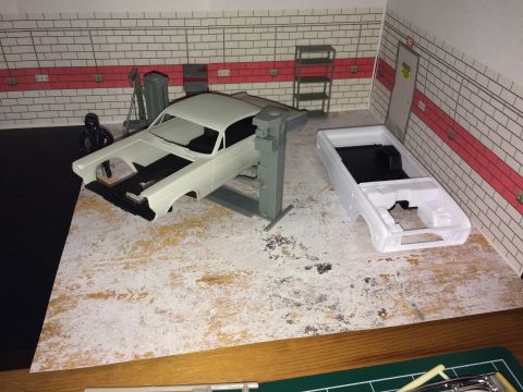 1989 Ford Bronco - A model of a white car body is on a lift in a diorama garage. Another white car body is on the floor next to it, and a figure in a black su…