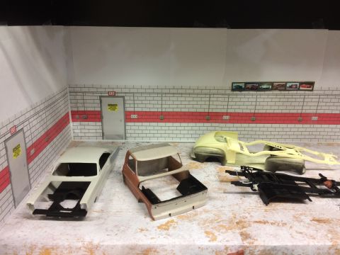 1989 Ford Bronco - A model garage scene with several unfinished model car bodies and chassis laid out on the floor. Two doors with caution signs are visible o…
