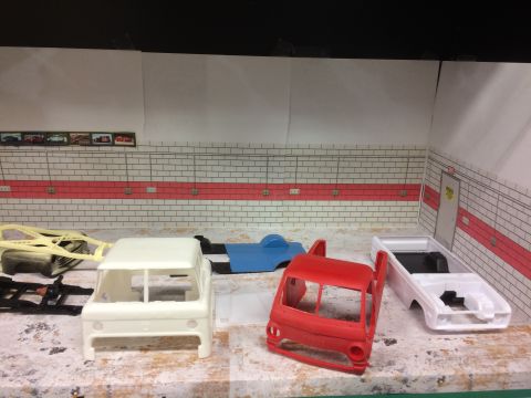 1989 Ford Bronco - A collection of unpainted plastic model vehicle parts are laid out on a surface in front of a diorama depicting a garage interior. Visible…