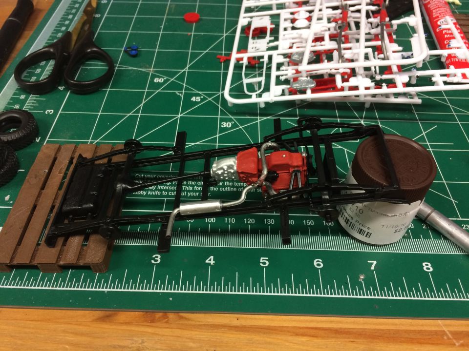 Model car chassis with engine and exhaust on a wooden platform on a cutting mat.