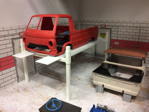 1989 Ford Bronco - A red model truck body sits on a white car lift in a diorama garage. A second, unpainted model truck body with a brown roof is parked to th…
