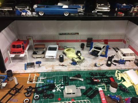 1989 Ford Bronco - A diorama depicting a model car repair shop with various car bodies, chassis, engines, and tools scattered around. A red pickup truck body…
