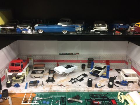 1989 Ford Bronco - A model car garage scene displays various unfinished car models and parts on shelves and a workbench. A red pickup truck model is elevated…
