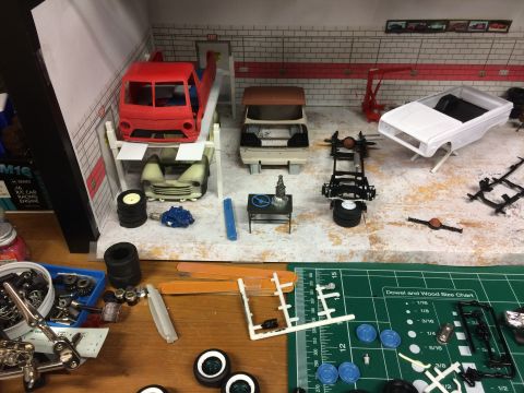 1989 Ford Bronco - A model car garage scene with various car parts and partially assembled model vehicles, including a red pickup truck on a lift and a white…