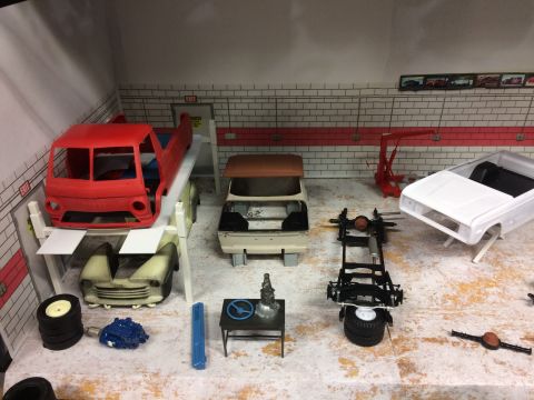 1989 Ford Bronco - A miniature garage scene displays several unfinished model car bodies and parts, including a red truck cab on a lift and a white pickup tru…