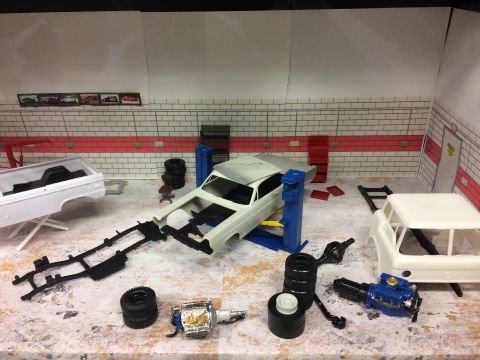 1989 Ford Bronco - A model car garage scene with a partially assembled white pickup truck body on a stand, a car chassis on a lift, and various engine and tir…