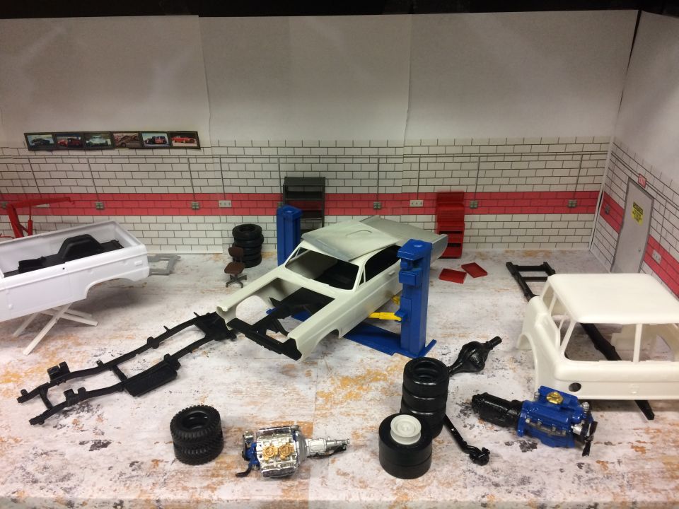 Model car garage scene with white pickup truck body, car chassis on lift, engine, and tires.