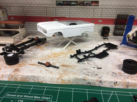 1989 Ford Bronco - A model of a white Ford Bronco body sits on a stand next to its chassis and other parts in a diorama of a garage. A second, partially assem…