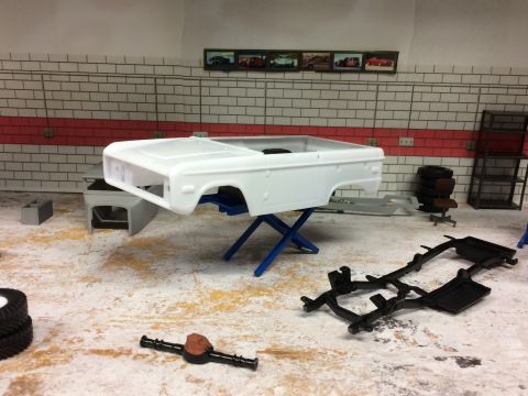 1989 Ford Bronco - A white model car body for a 1989 Ford Bronco is displayed on a blue scissor lift in a diorama garage setting. A black model frame and a di…