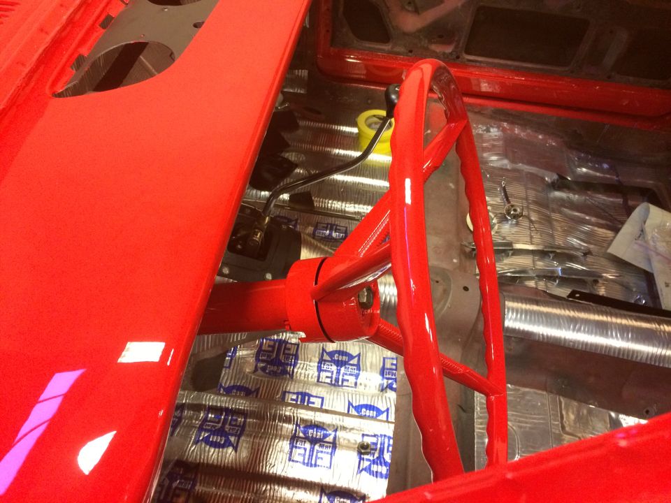 Red steering wheel and column in a classic car interior with sound deadening on the floor.