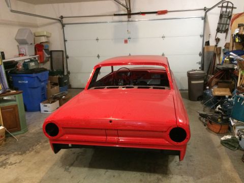 1964 Ford Fairlane - A bright red vintage car body sits in a garage, facing forward. The car is missing its headlights and has exposed interior components.