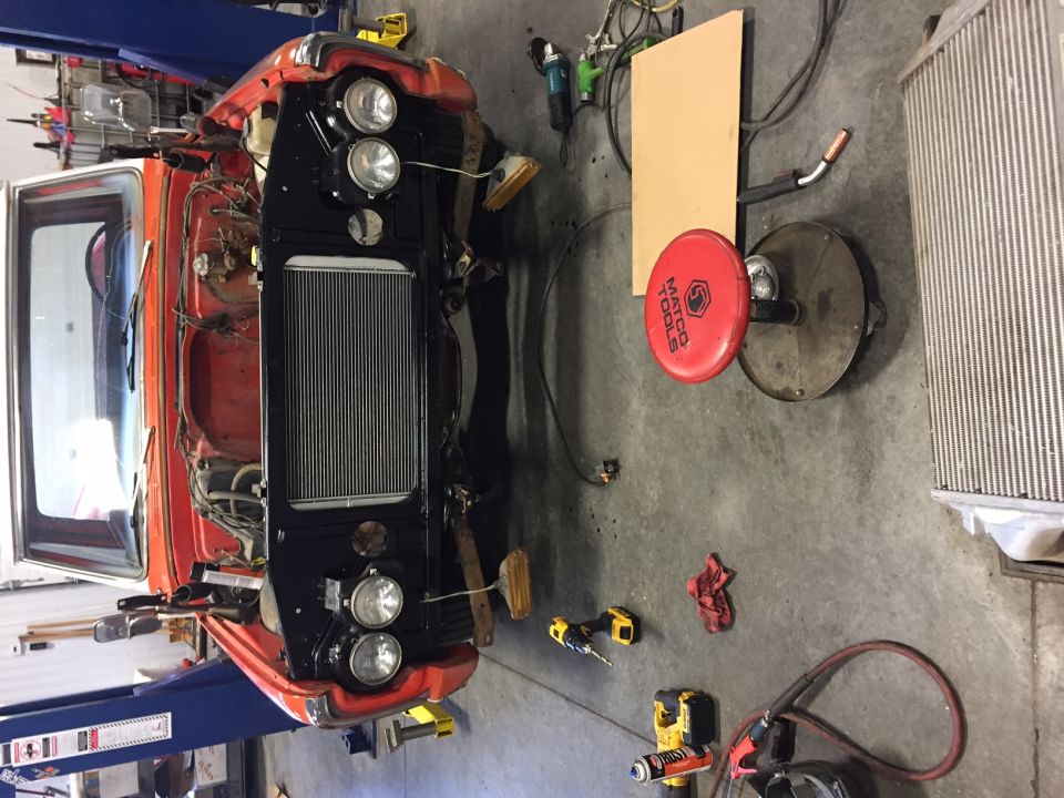 Disassembled front of an orange and white vehicle on a lift, showing engine bay components and tools on the floor.