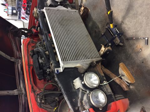 1984 Ford Bronco - A red vehicle's engine bay is shown with the hood open, revealing a large radiator and various engine components. A blue hydraulic jack and…