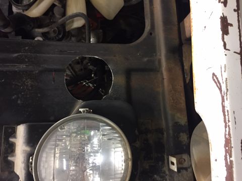 1984 Ford Bronco - A circular hole has been cut into the black metal panel of a vehicle, revealing engine components and wiring behind it. A headlight assembl…