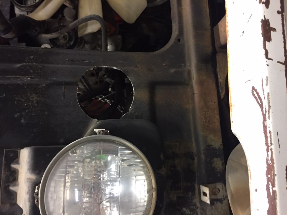 Close-up of a hole cut in a vehicle's black metal panel, with engine parts and wiring visible, and a headlight below.
