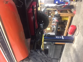 1984 Ford Bronco - The underside of an orange vehicle is shown, lifted on a hydraulic lift. Visible components include a large fuel tank, suspension parts, an…