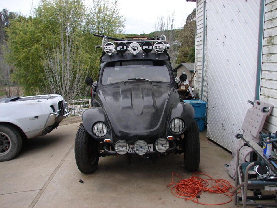 Modified black Volkswagen Beetle with roof rack and lights, parked next to a white building.