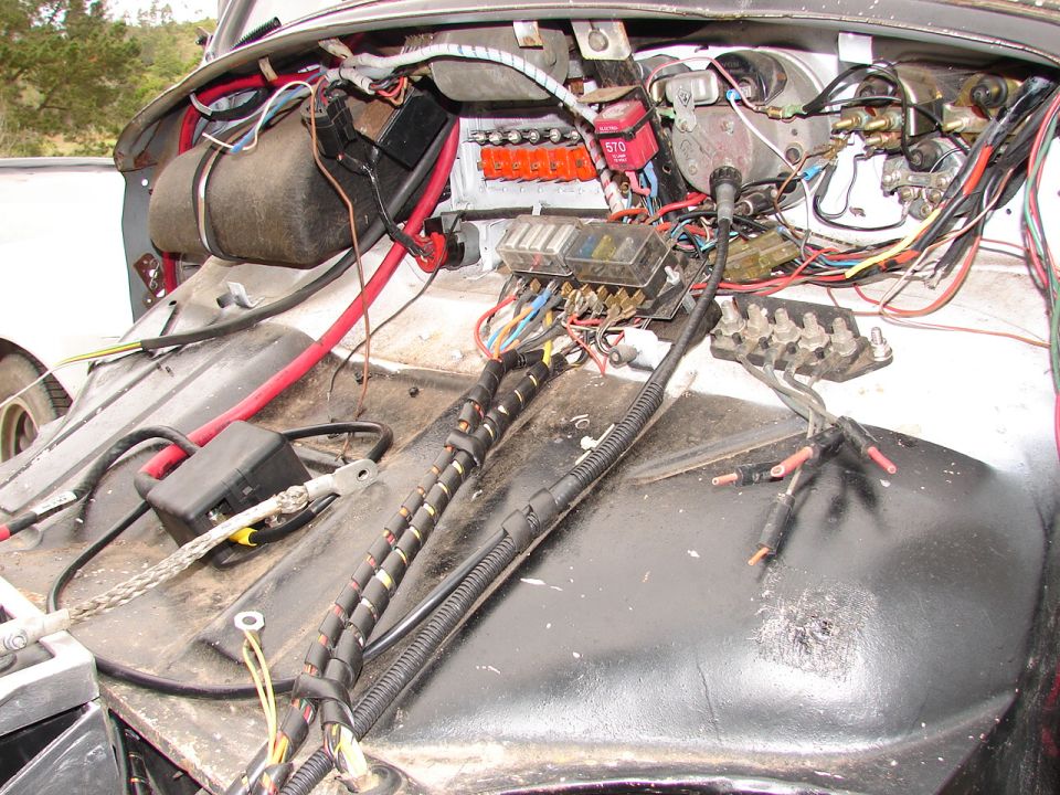 Close-up of a classic car engine bay showing wiring, fuse boxes, and battery.