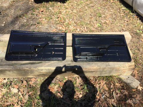 1979 Ford Bronco - Two black plastic interior door panels are shown resting on wooden beams outdoors. The panels have a textured upper section and a molded ha…