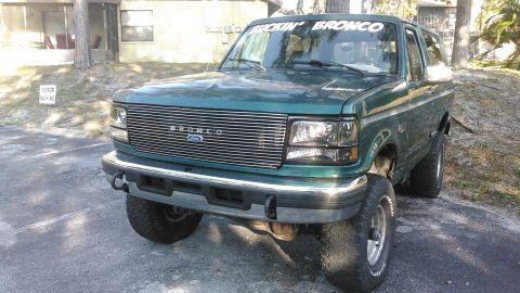 1996 Ford Bronco - A front, slightly low-angle view of a dark green Ford Bronco parked on asphalt. A white banner reading "BUCKIN' BRONCO" is visible across t…