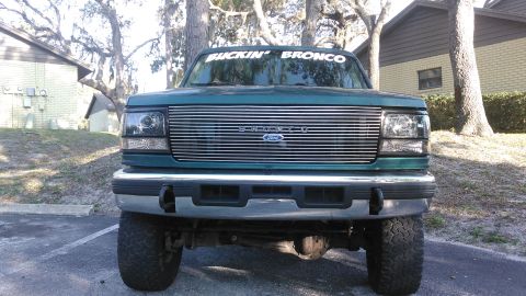 1996 Ford Bronco - Front view of a dark teal 1996 Ford Bronco with a chrome grille and a white banner on the windshield that reads "BUCKIN' BRONCO". The vehic…