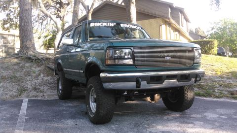 1996 Ford Bronco - A dark green 1996 Ford Bronco is parked in a lot with a white banner on the windshield that reads "BUCKIN' BRONCO". The vehicle is shown fr…