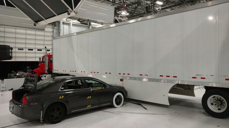 Dark sedan with yellow impact sensors is parked next to a white semi-trailer. The sedan's trunk is open