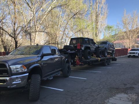1991 Ford Bronco - A Ford F-350 Super Duty tows a trailer carrying a Jeep Wrangler and a Ford Bronco, with another white truck parked nearby.