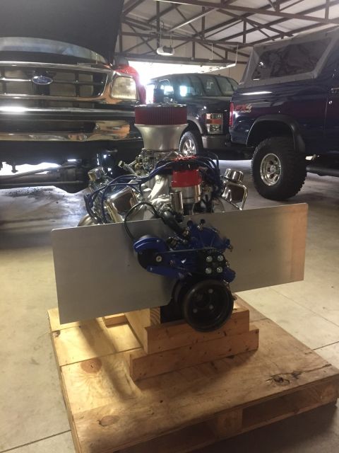 1982 Ford Bronco - A custom V8 engine with a blue intake manifold and red air filter sits on a wooden pallet in a garage. The engine is in front of a dark blu…