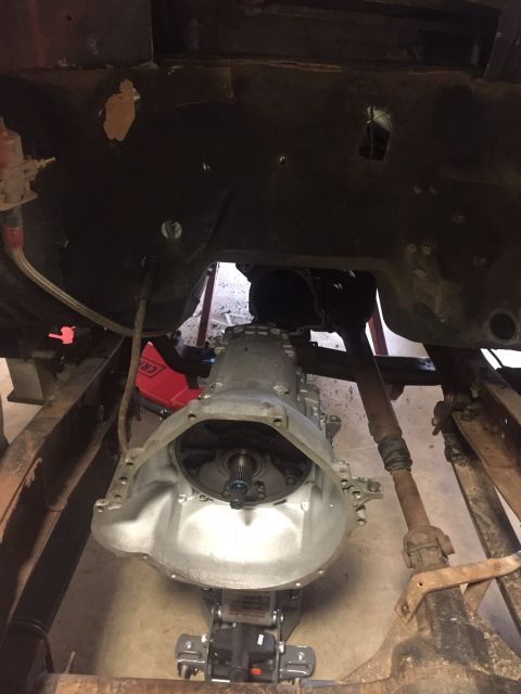 1982 Ford Bronco - A silver automatic transmission is supported by a hydraulic jack beneath the frame of a vehicle. The transmission bell housing is visible,…