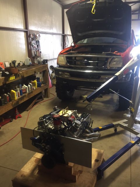 1982 Ford Bronco - A V8 engine is mounted on a stand in front of a Ford truck with its hood open. The engine appears to be a custom build with chrome valve co…