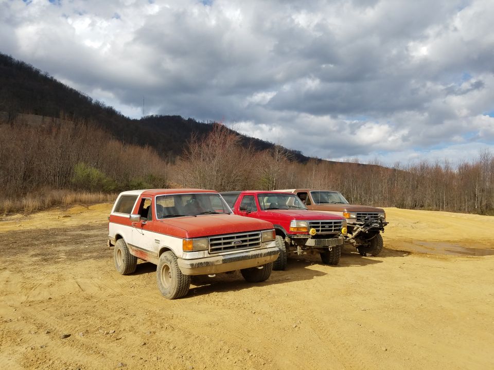 Three vintage Ford trucks parked on a dirt lot. The lead vehicle is a red and white Bronco.
